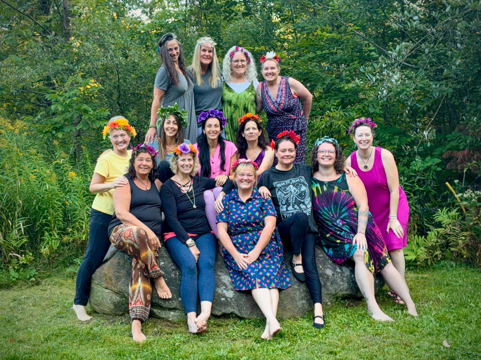 A group of Held Sacred sisters wearing flower crowns gathered around a boulder in the woods