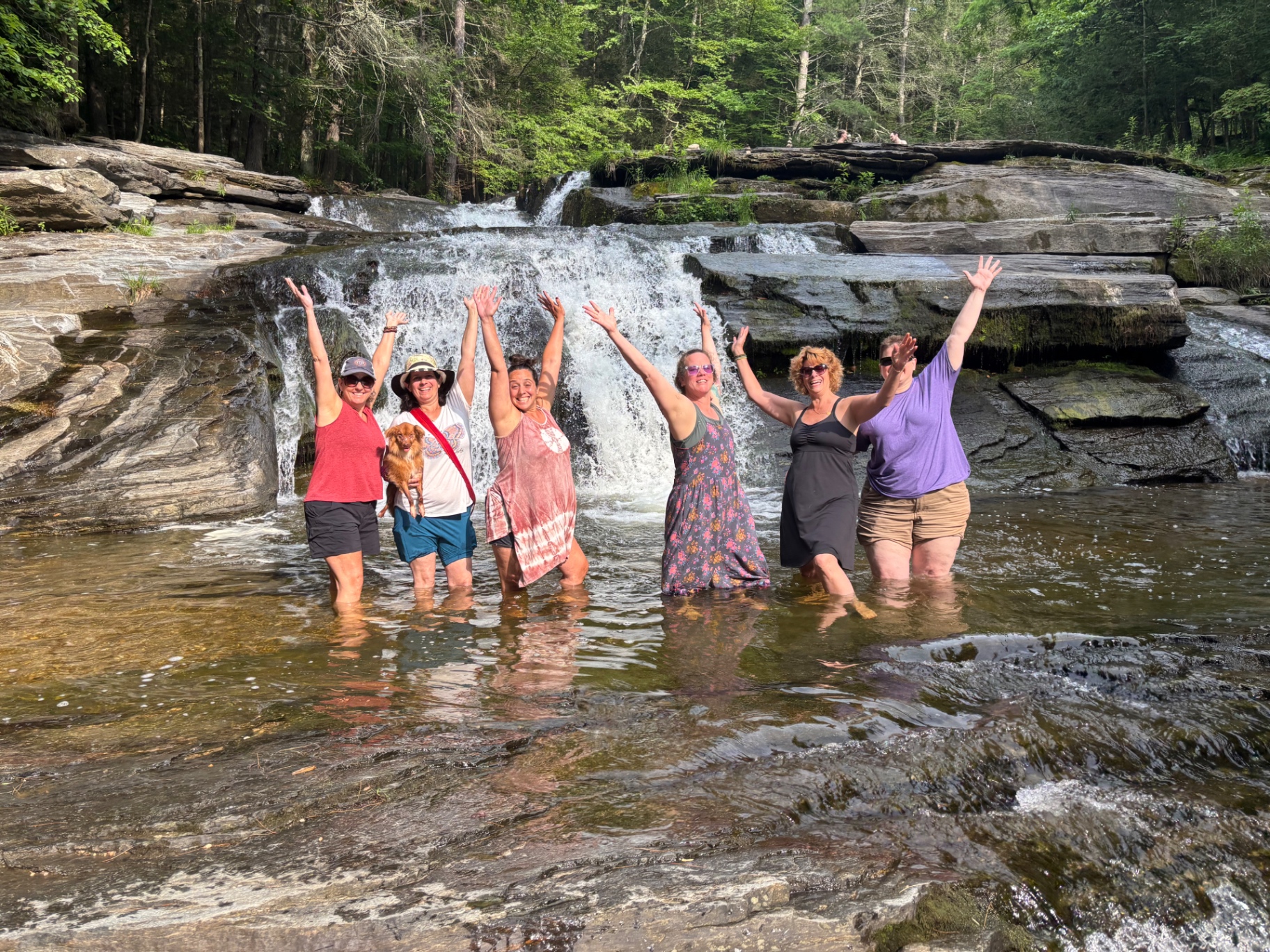 Held Sacred sisters standing in a river with arms raised in front of a waterfall